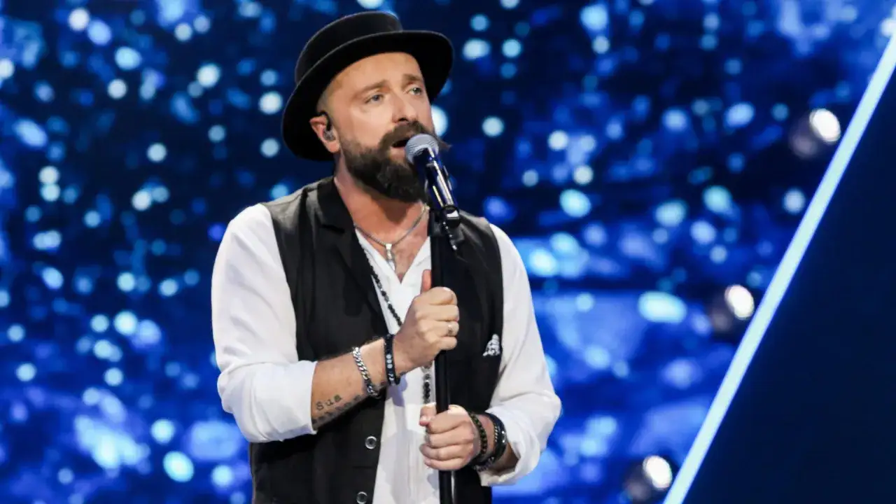 Łukasz Drapała w czarnym kapeluszu śpiewa na scenie, tworząc szalony bal dźwięków.