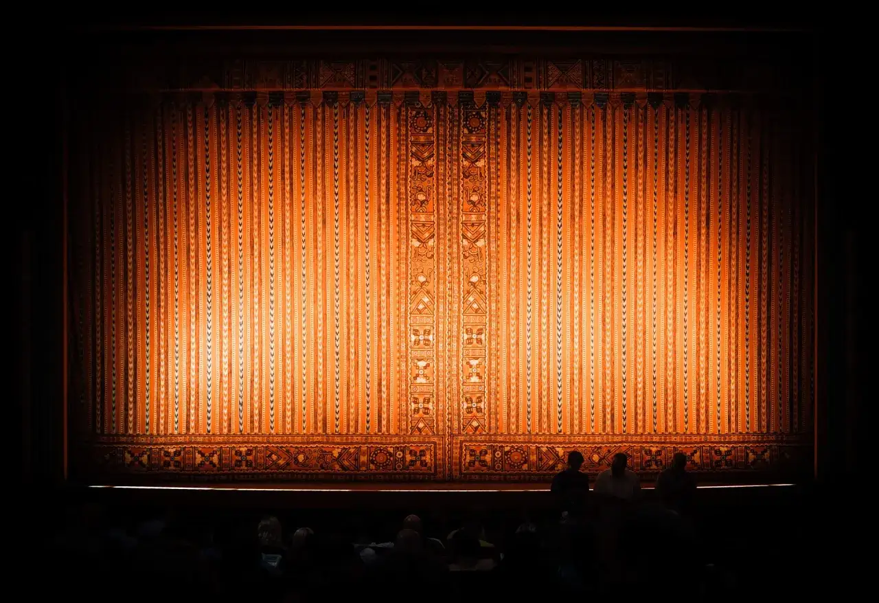 Złocista kurtyna teatralna, ozdobiona wzorami, czeka na rozpoczęcie spektaklu. Widownia w półmroku zastanawia się, co to będzie dalej.