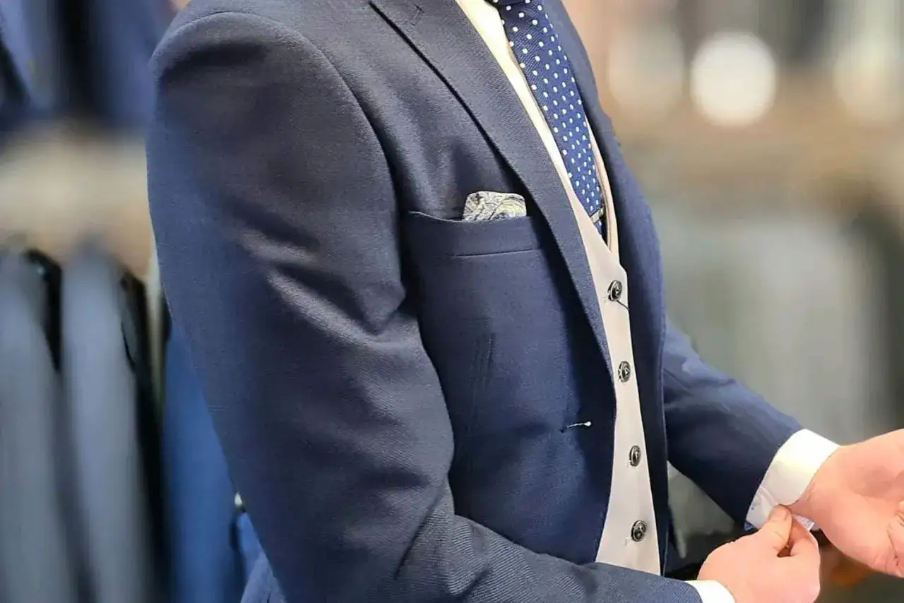A groom gets his suit fitted. He's wearing a navy blue suit jacket, a light grey vest, and a blue polka dot tie with a patterned pocket square.