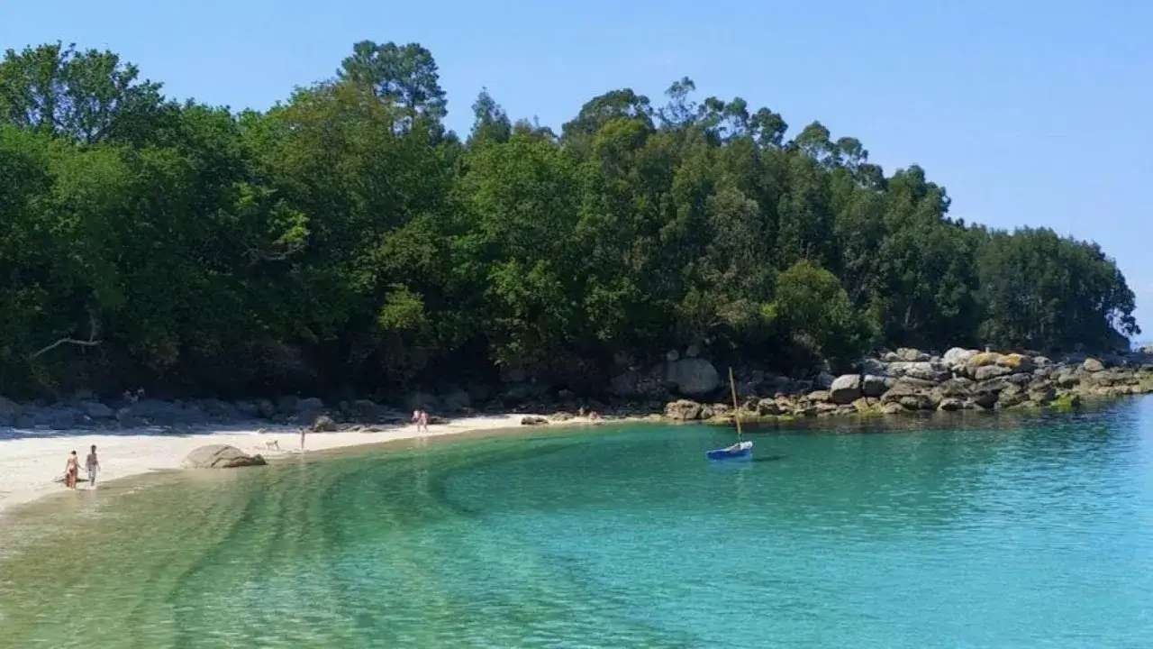 Aguas cristalinas y arena blanca en la playa de Vilari&ntilde;o, con un peque&ntilde;o velero meci&eacute;ndose suavemente. Un d&iacute;a perfecto para disfrutar del sol y la naturaleza.