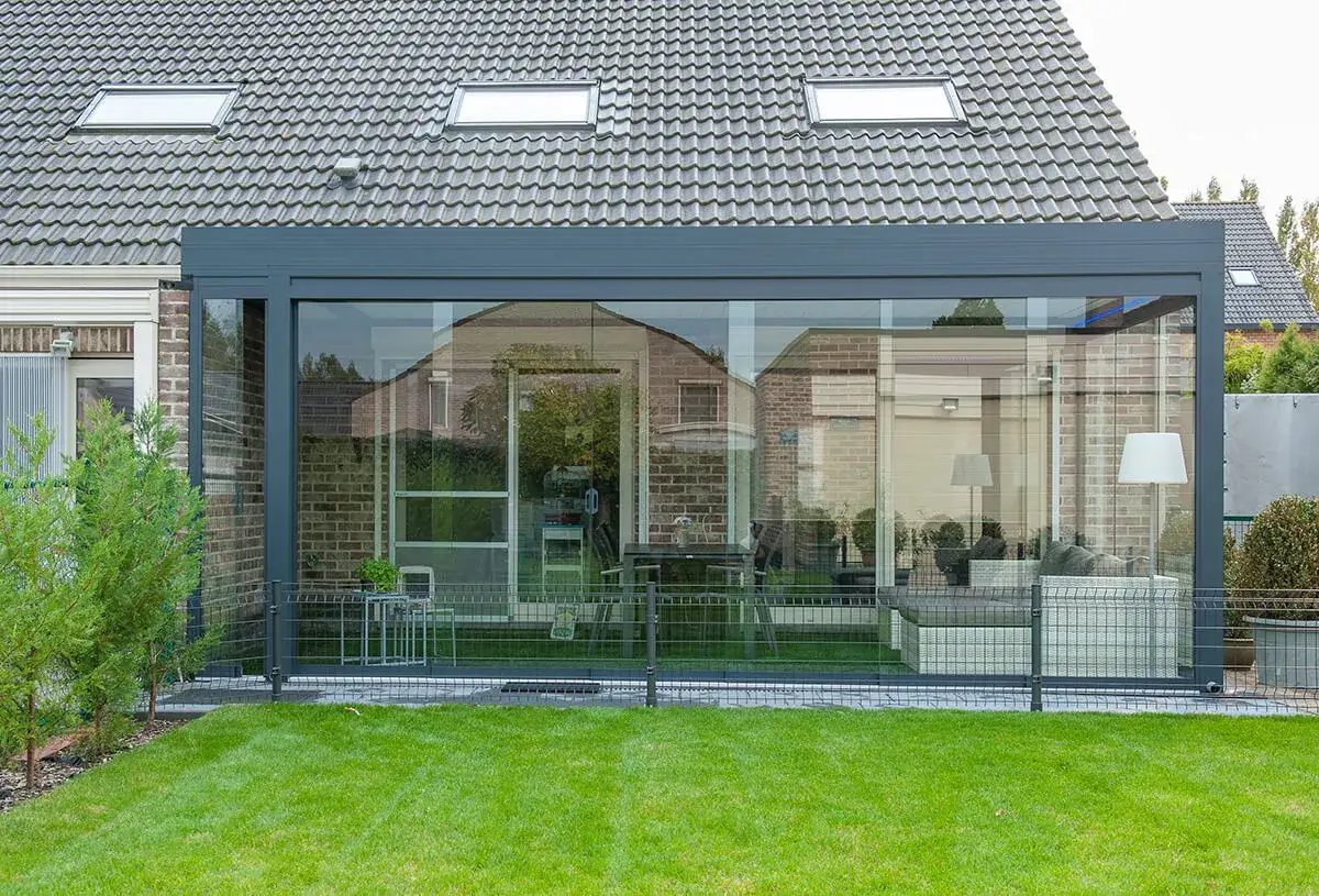 Une pergola fermer en polycarbonate avec des murs vitrés, offrant une vue sur un salon moderne et un jardin verdoyant.