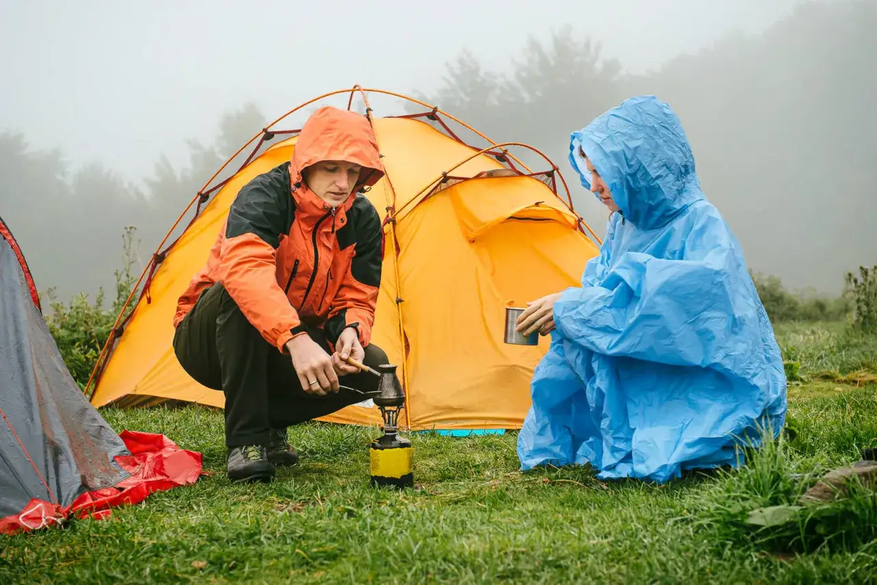 Zwei Personen in Regenponchos bereiten sich auf das Zelten bei Regen vor. Eine Person kocht auf einem Campingkocher, die andere h&auml;lt eine Tasse.