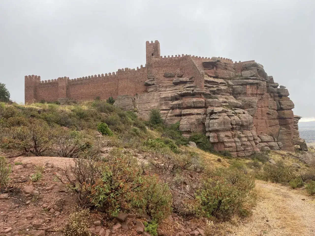 Imponente castillo de **Villar del Salz** coronando una formaci&oacute;n rocosa rojiza, con vegetaci&oacute;n baja y un cielo nublado.