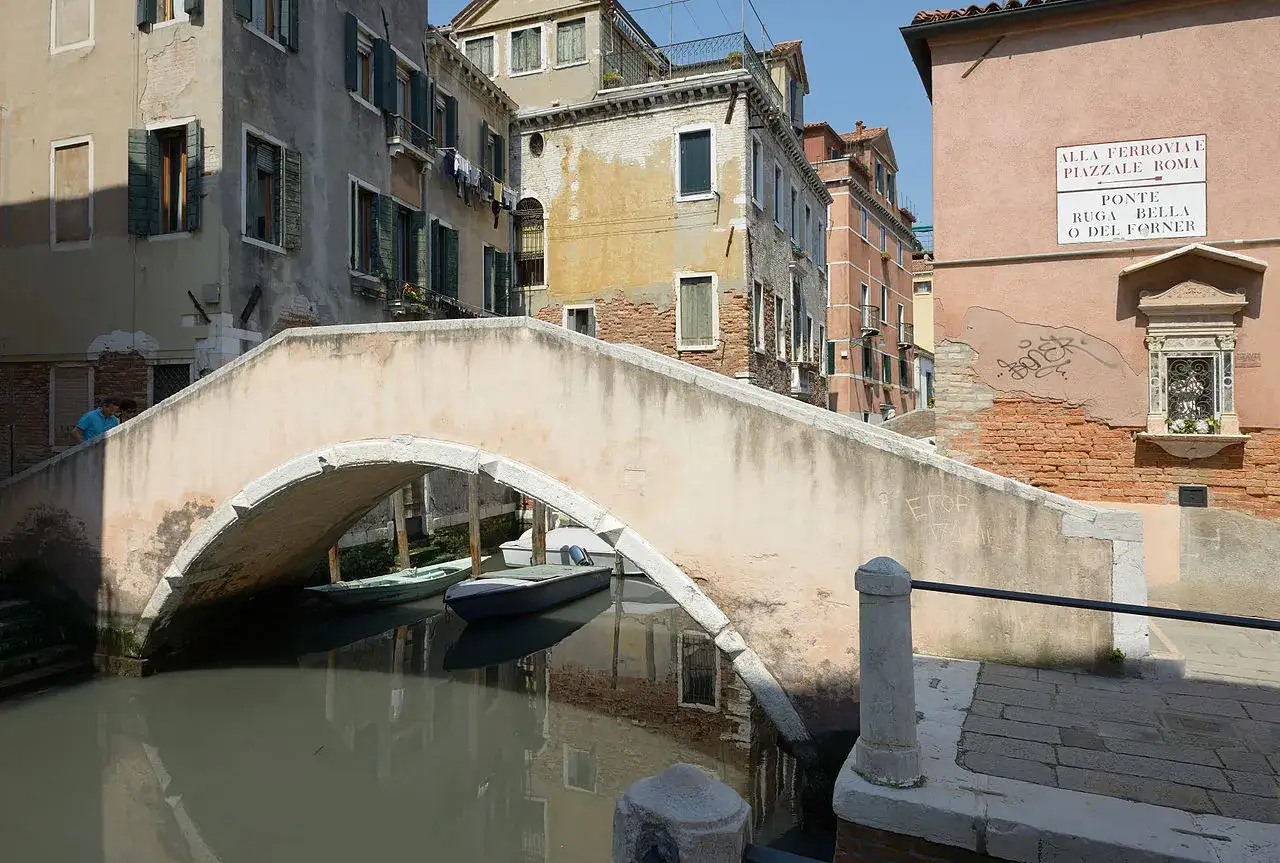 Un ponte ad arco in una stretta calle veneziana, con barche ormeggiate nel canale sottostante e palazzi storici che si affacciano.