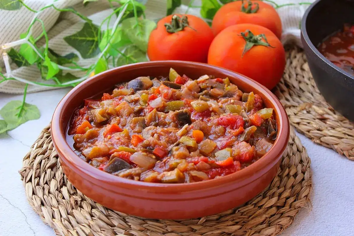 Un cuenco de barro rebosante de pisto murciano, con trozos de tomate, pimiento, berenjena y cebolla. Tomates frescos y hojas verdes decoran la escena.