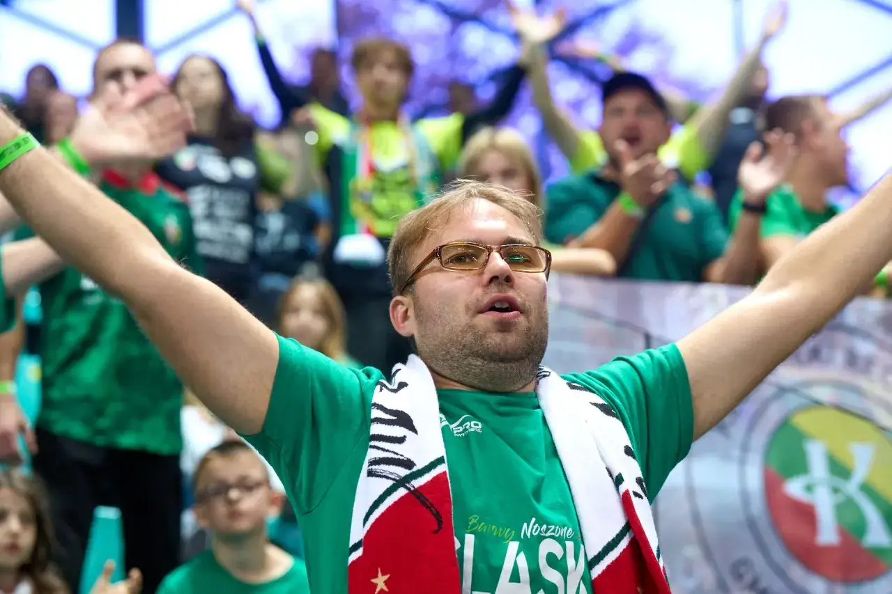 Mężczyzna w okularach z szalikiem kibicuje, w tle tłum w zielonych koszulkach. Industria Kielce!
