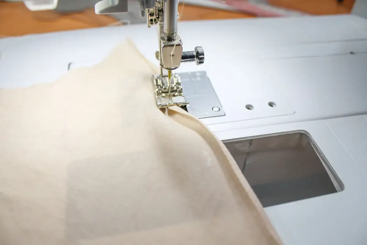 A sewing machine creates a narrow hem on a light beige fabric.