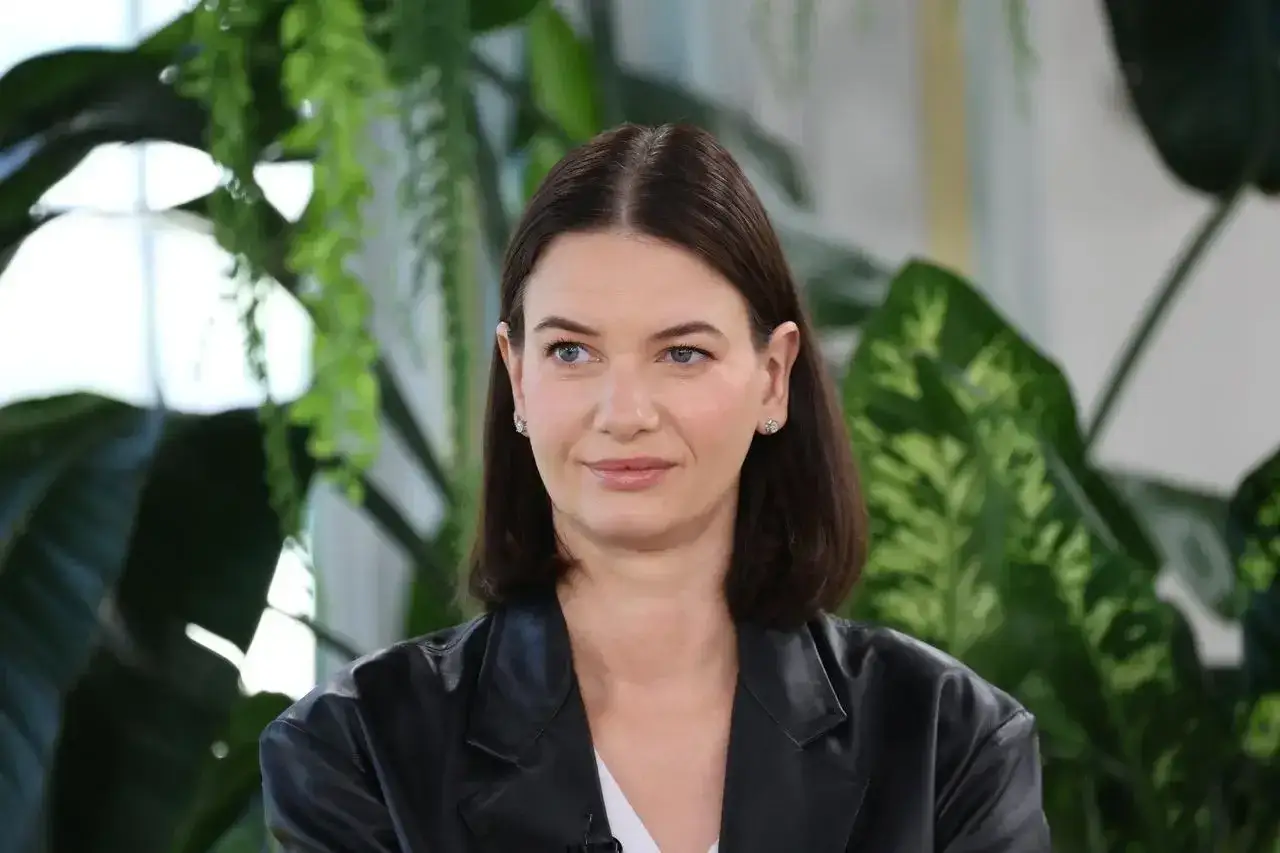 Karolina Malinowska Janiak w czarnej marynarce, z błękitnymi oczami, na tle zielonych roślin.