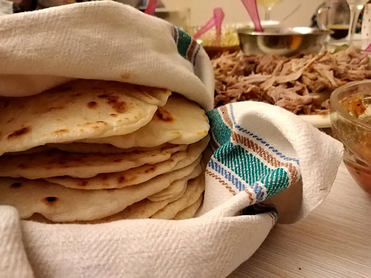 Stos świeżo upieczonych placków tortilla oryginalny przepis, gotowych do podania z szarpaną wieprzowiną i dodatkami.