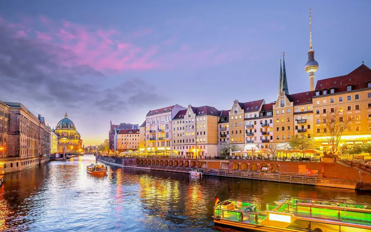En vacker kv&auml;ll vid floden i Berlin, med upplysta byggnader och en b&aring;t som passerar. Perfekt f&ouml;r kv&auml;llsaktiviteter i Berlin.