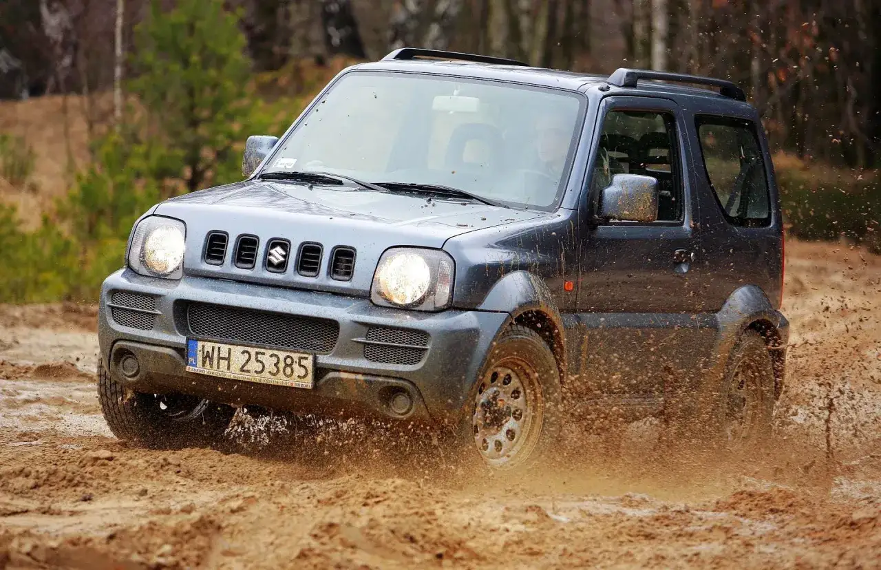 Szary Suzuki Jimny (1998) z 1998 roku pokonuje błotniste wyzwanie, rozpryskując błoto na boki.