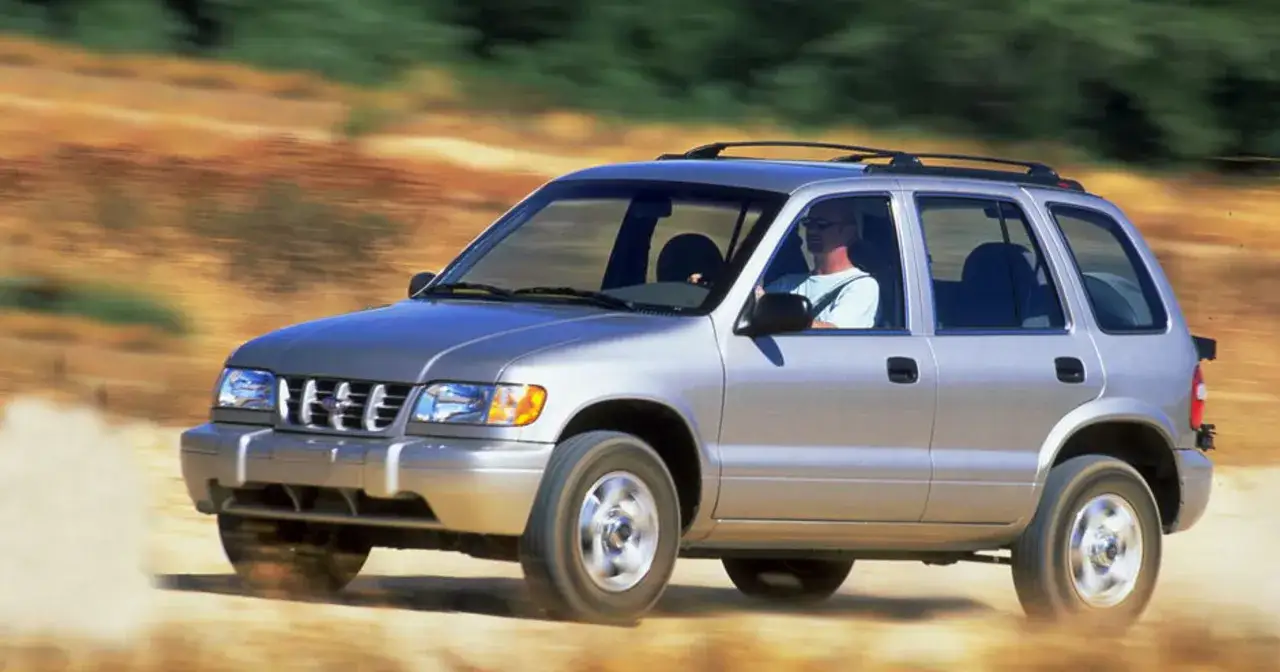 Srebrny Kia Sportage 1 z mężczyzną za kierownicą jedzie po piaszczystej drodze, wzbijając kurz.