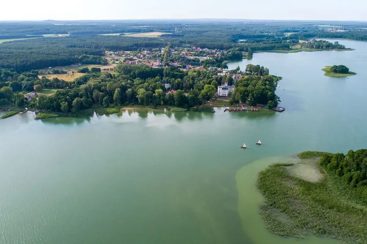 Malownicze jezioro Niesłysz z dwoma żaglówkami, otoczone lasami i zabudowaniami.