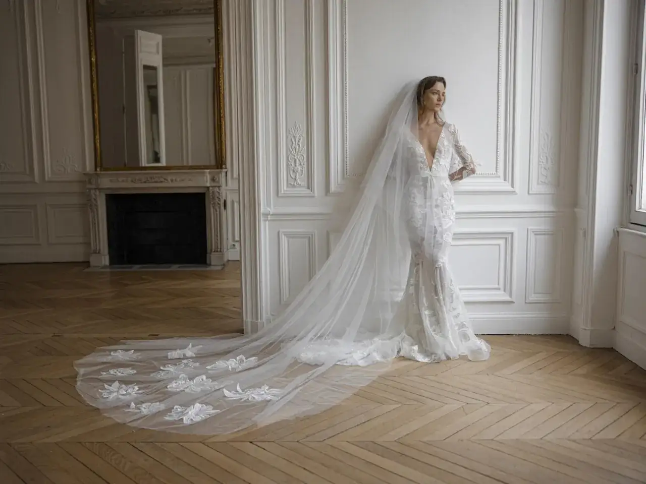 Braut in einem Spitzenkleid mit tiefem Ausschnitt und einem langen, bestickten Schleier, der elegant über den Holzboden fällt. Perfekt für den **Schleier Standesamt**.