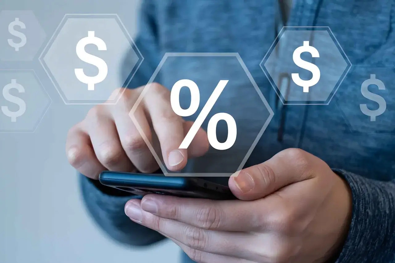 Mani che scorrono su uno smartphone con icone di dollari e percentuali. Scopri cosa copre l'assicurazione annullamento viaggio per viaggiare sereni.