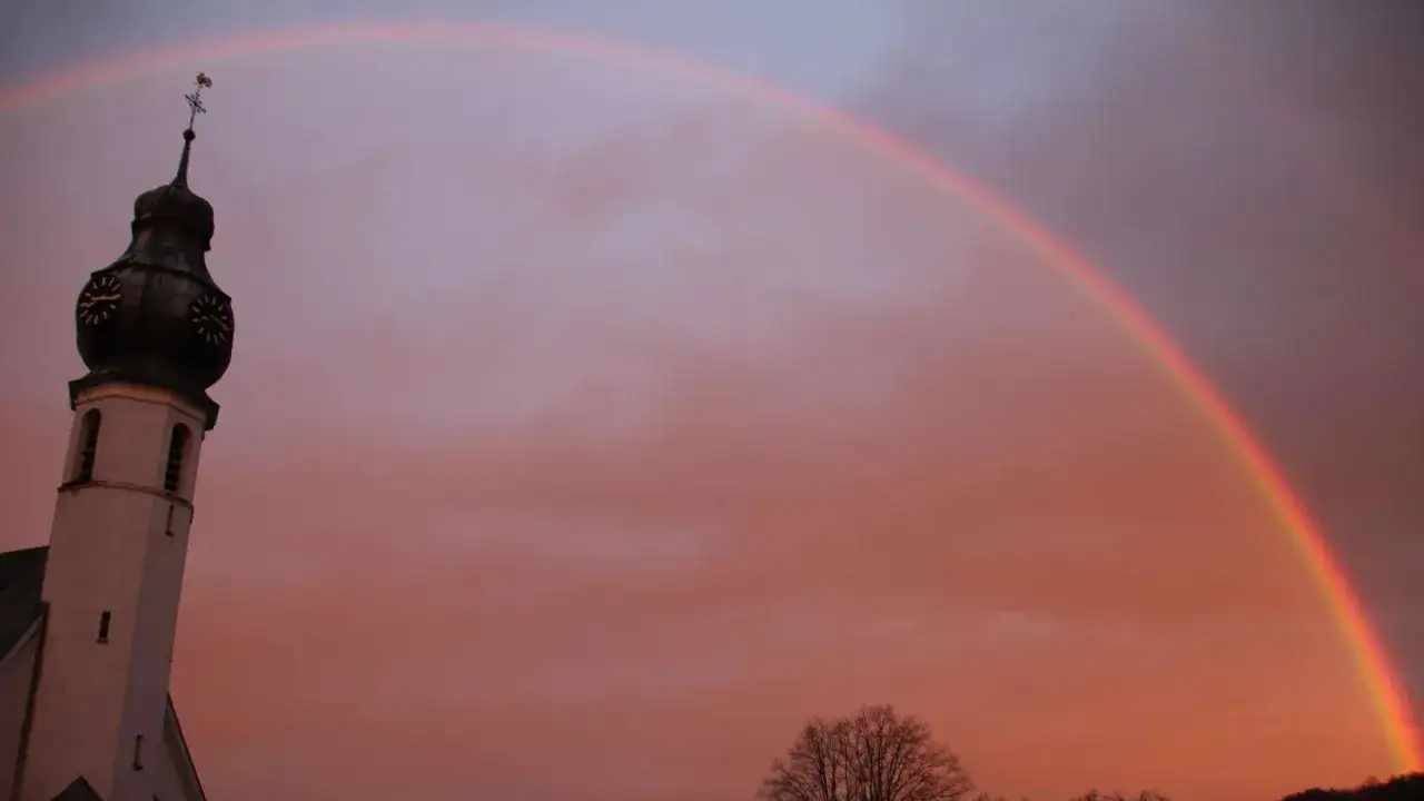 Kirchturm im Abendrot mit einem Regenbogen, der Hoffnung und Neuanfang symbolisiert.
