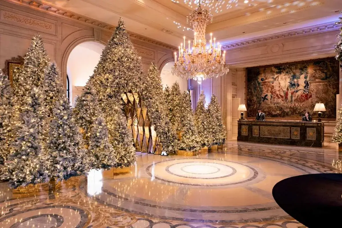 Un hall d'h&ocirc;tel somptueux, d&eacute;cor&eacute; de nombreux sapins illumin&eacute;s pour une **soir&eacute;e de No&euml;l** f&eacute;erique. Un lustre scintille au plafond.
