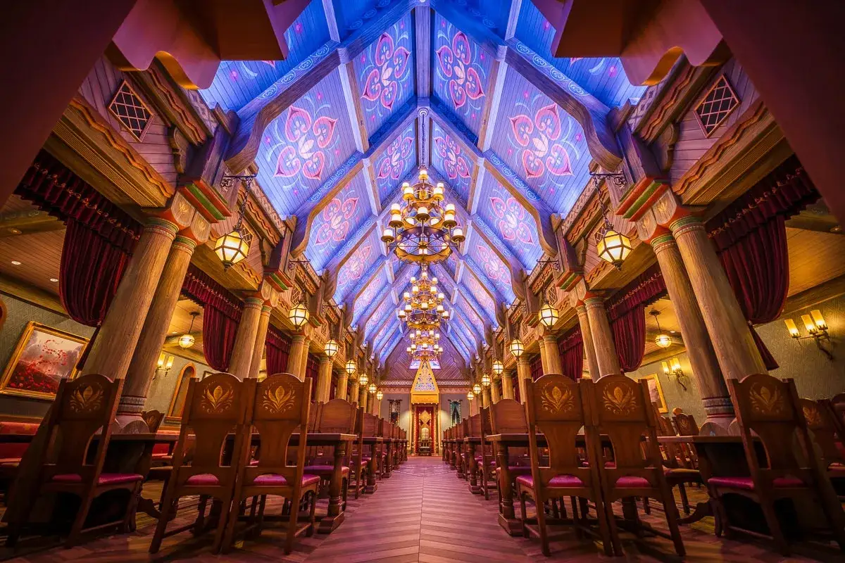 Ein prunkvoller Speisesaal im Stil von **Frozen Disneyland**, mit Holzs&auml;ulen, Kronleuchtern und einem blau-lila beleuchteten Dach mit Ornamenten.