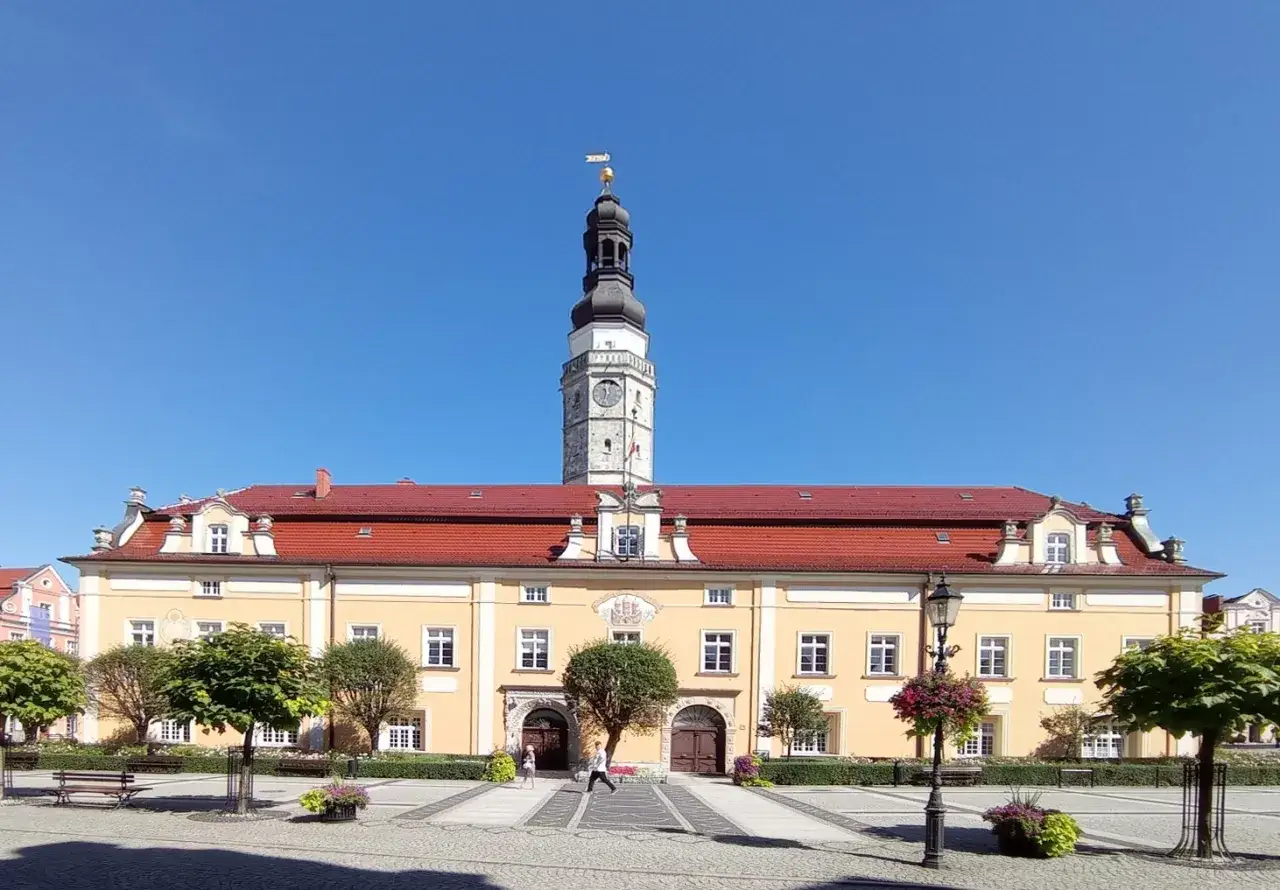 Słoneczny rynek w Bolesławcu z żółtym ratuszem i wieżą zegarową. Spacerują ludzie, rosną drzewka i kwitną kwiaty.