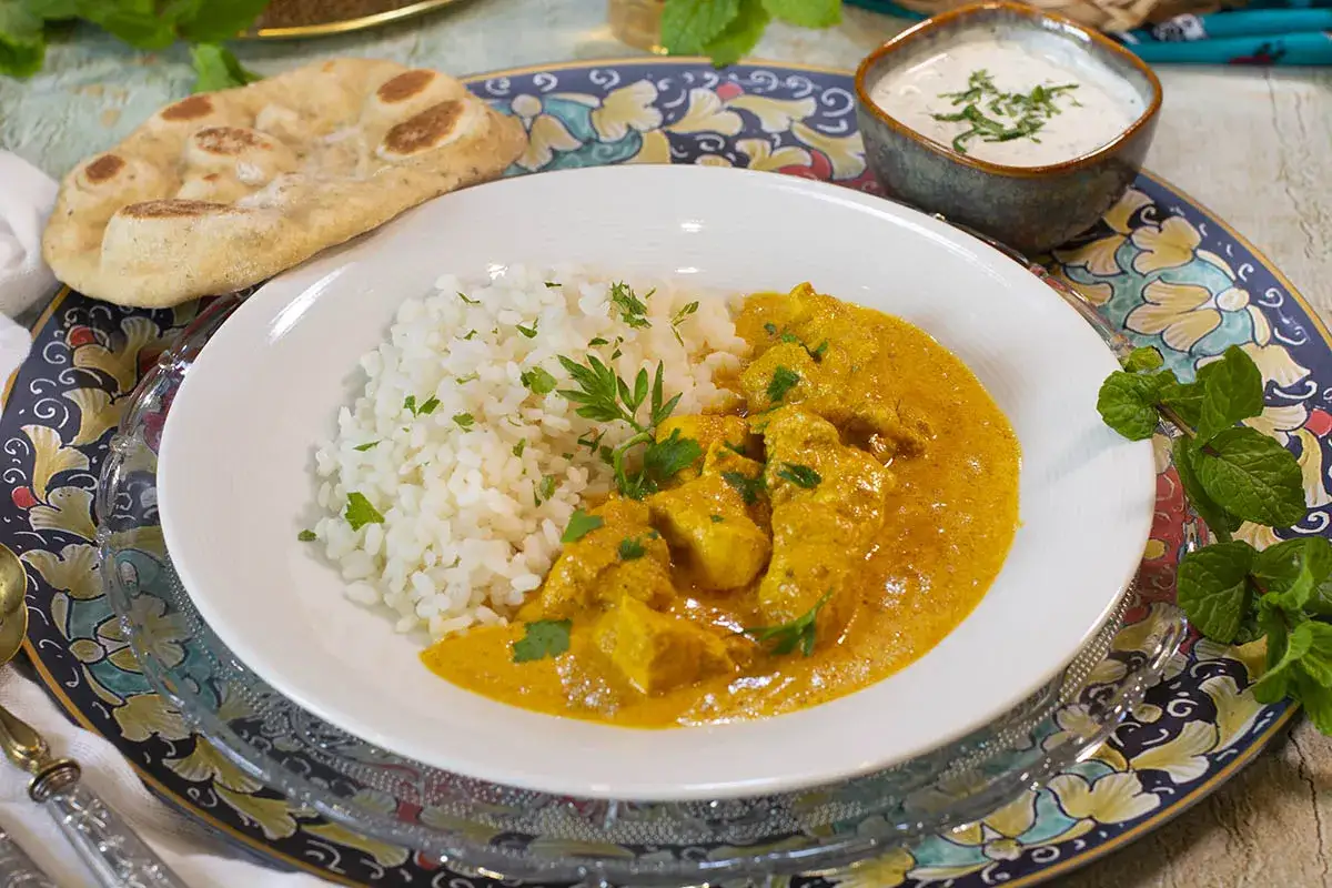 Delicioso pollo tikka masala cremoso con arroz blanco y pan naan, acompañado de una salsa de yogur y menta fresca.