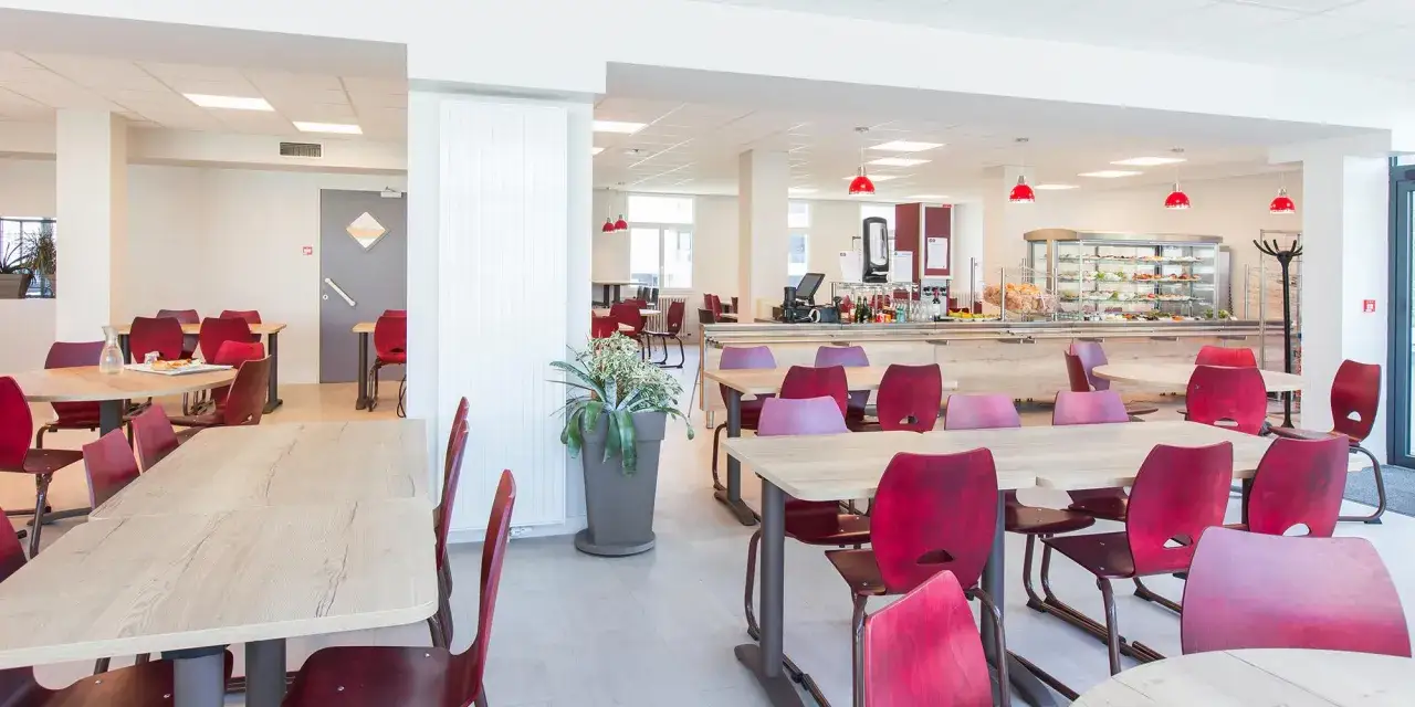 La timonerie Saint-Malo : une cafétéria lumineuse avec tables en bois clair et chaises rouges, un buffet garni et des plantes vertes.