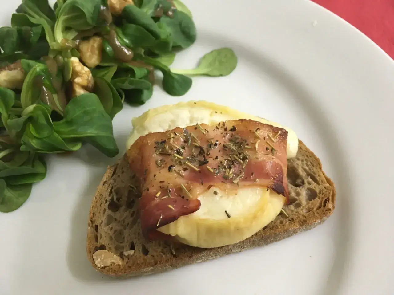 Tartine de pain noir avec ch&egrave;vre chaud, lard et herbes, accompagn&eacute;e d'une salade verte aux noix.