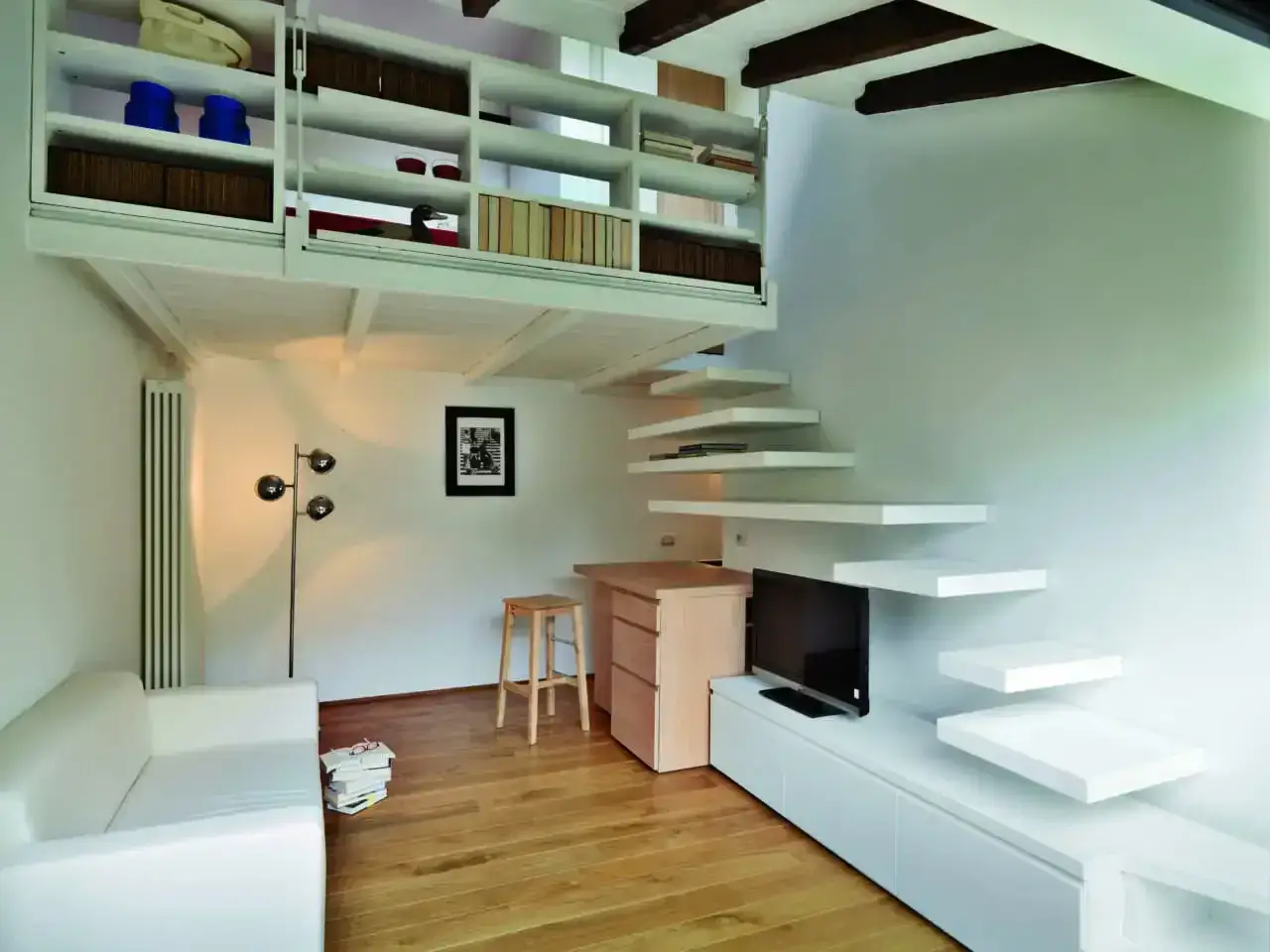 Open space loft con soppalco. Soggiorno moderno con divano bianco, libreria a vista e scala sospesa che conduce al soppalco.