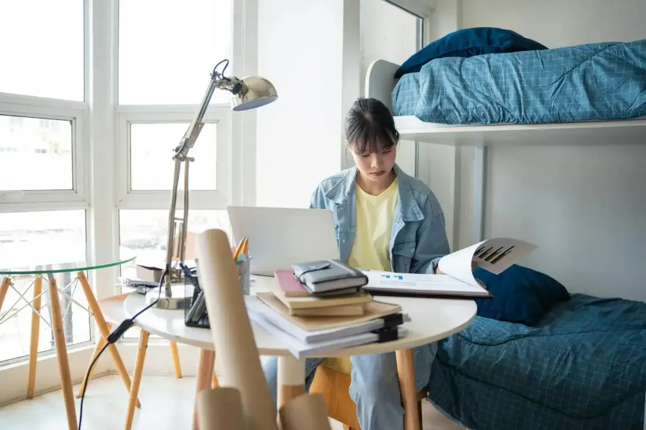 Jeune femme travaillant sur son ordinateur portable dans une chambre d'&eacute;tudiant &agrave; Singapour, avec un lit superpos&eacute; bleu en arri&egrave;re-plan.