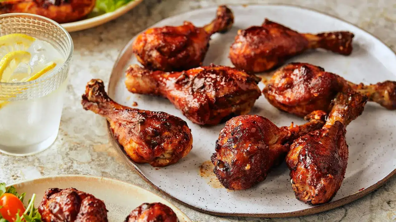 Plusieurs pilons de poulet grillés, brillants de sauce barbecue, sont disposés sur une assiette. Un verre d'eau glacée avec des rondelles de citron est à côté.