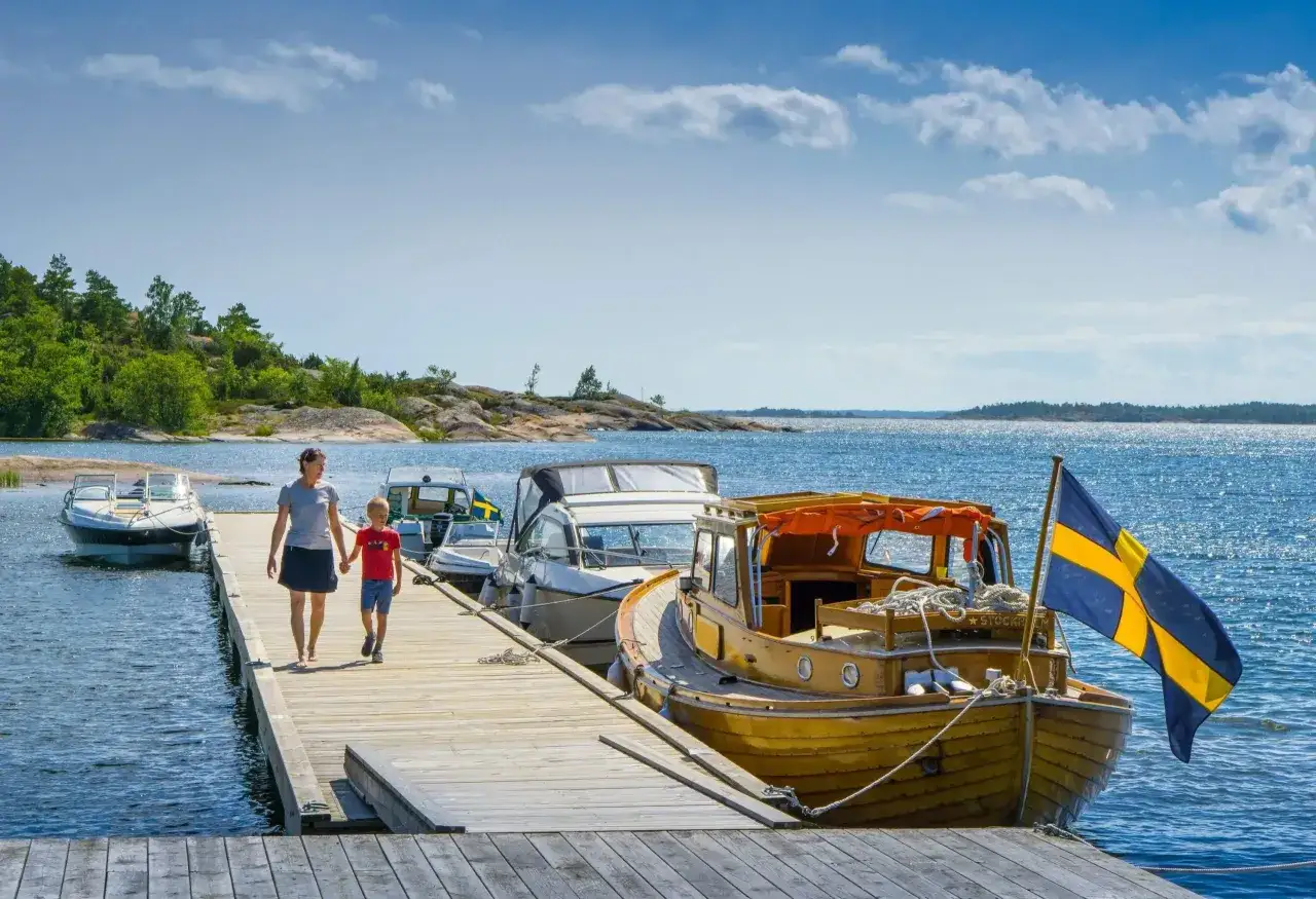 En kvinna och ett barn går på en brygga med båtar. En svensk flagga vajar. En perfekt semester i Sverige med barn.