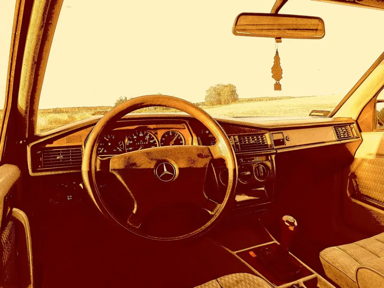 Wnętrze klasycznego Mercedes 190e. Kierownica, deska rozdzielcza i siedzenie pasażera. Za oknem pole i drzewa.
