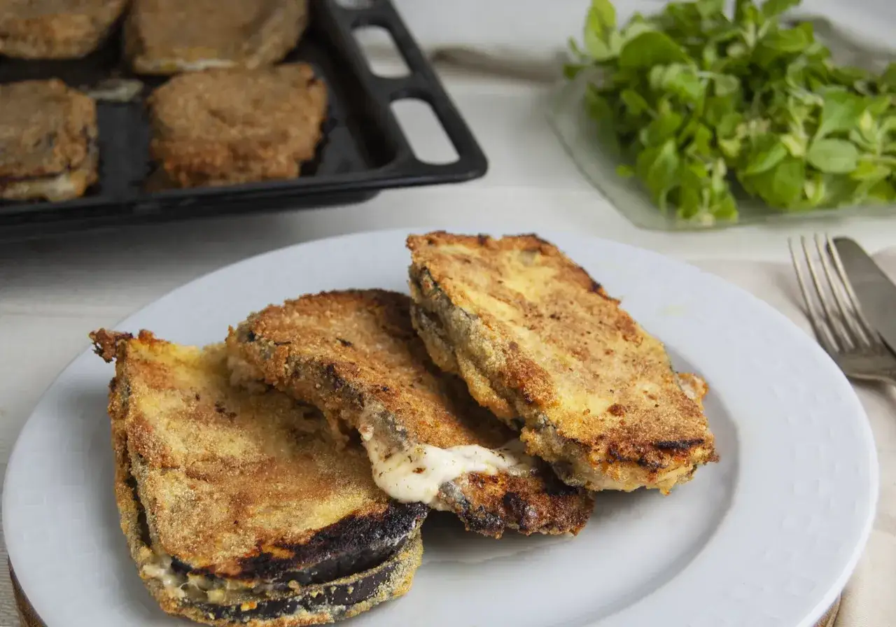 Tre cordon bleu di melanzane dorati e filanti, serviti su un piatto bianco, con un contorno di insalata fresca e una teglia sullo sfondo.