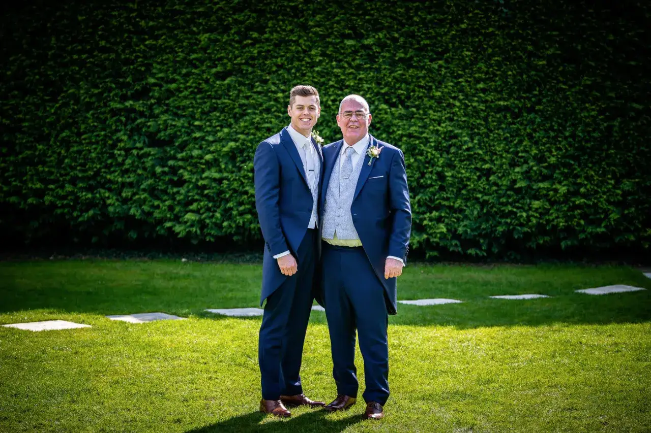 Two men in matching navy blue tailcoats, one older, the father of the groom, looking proud.