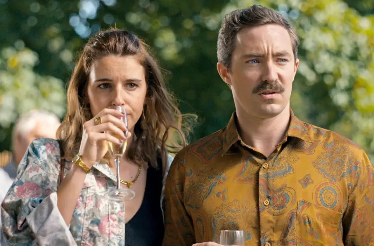 Ein Mann mit Schnurrbart und eine Frau mit Champagnerglas, beide in bunten Hemden, scheinen in der zweiten Staffel von