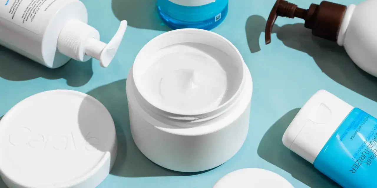 En öppen burk med vit body lotion i ansiktet, omgiven av andra hudvårdsprodukter.