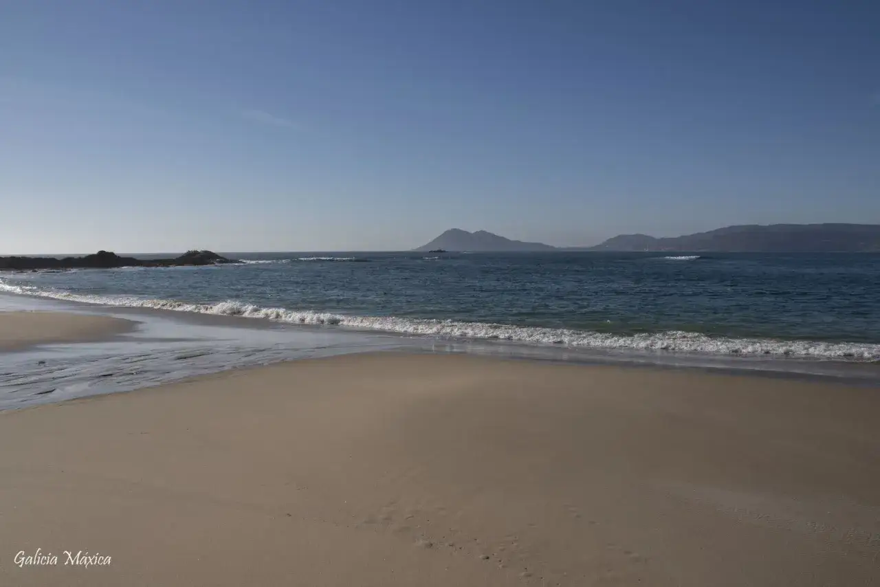 Arena dorada y mar azul en la playa de Cabeiro, con rocas a la izquierda y monta&ntilde;as en el horizonte.