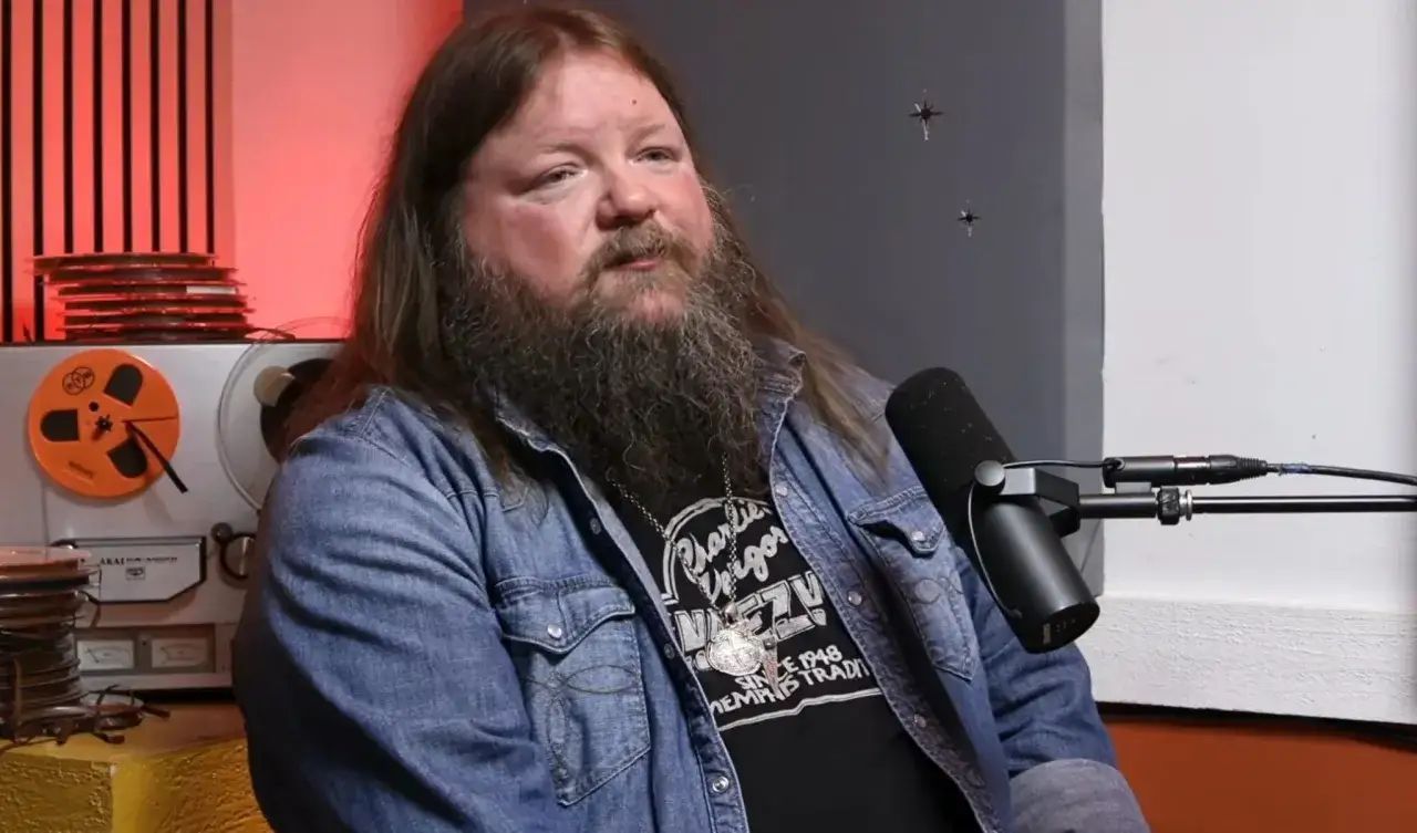 A man with a long beard and hair, wearing a denim shirt over a t-shirt with 