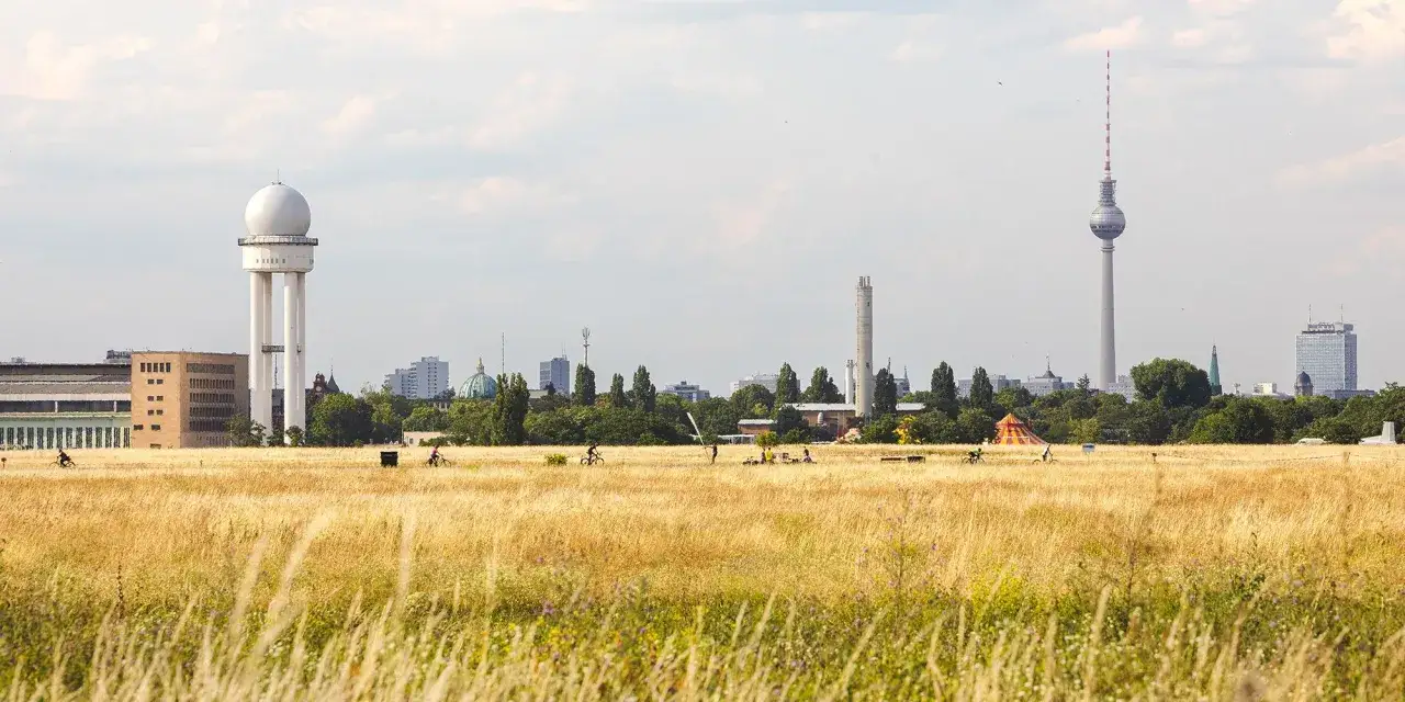 Ein weites Feld mit hohem Gras und Radfahrern, im Hintergrund die Skyline Berlins mit dem Fernsehturm und dem alten Radarturm. Ein besonderes in Berlin.