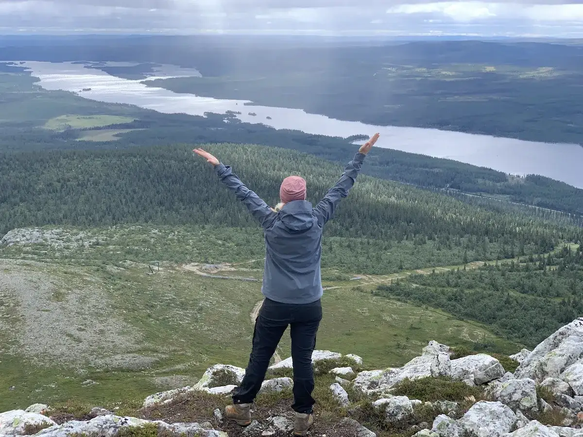 En person med armarna uppstr&auml;ckta njuter av utsikten &ouml;ver en sj&ouml; och skog under en vandring vid Ramundberget.