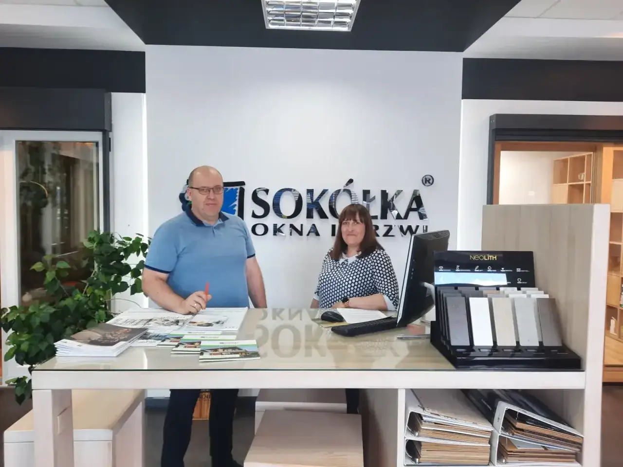 Para pracowników w salonie okien i drzwi, z logo firmy na ścianie.