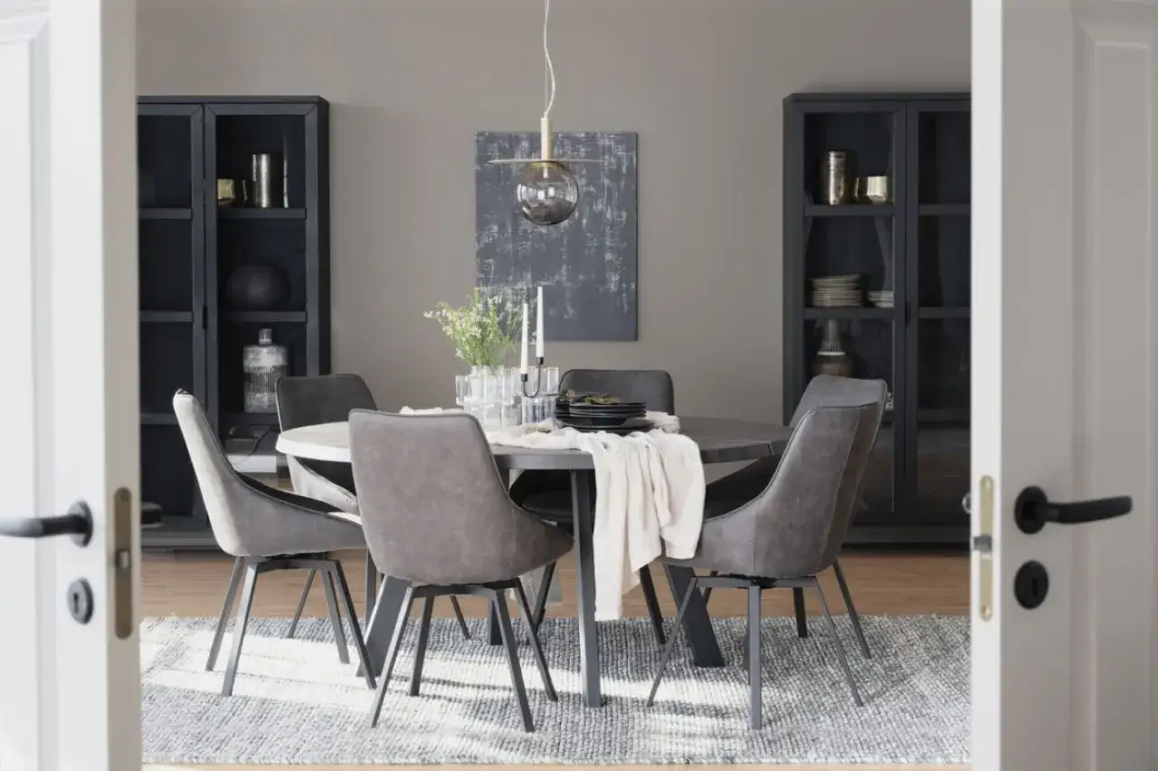 Elegant dukning runt ett runt bord med gr&aring; stolar. En vit duk med ljus och blommor skapar en inbjudande atmosf&auml;r.