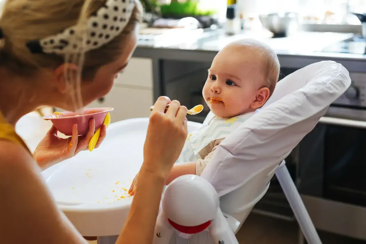 Mama karmi dziecko łyżeczką, rozpoczynając rozszerzanie diety. Maluch ma pomarańczowe jedzenie na buzi.