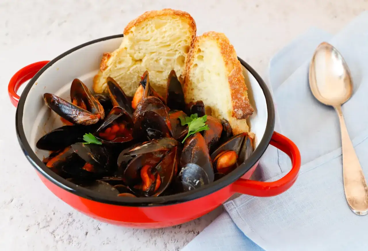 Un invitante guazzetto di cozze in una pentola rossa, accompagnato da pane croccante e un cucchiaio d'argento.