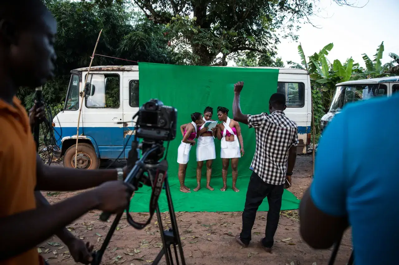 Ekipa filmowa tworzy scenę do filmu w stylu nollywood. Trzy kobiety w strojach ludowych stoją przed zielonym ekranem, a reżyser kieruje ujęciem.