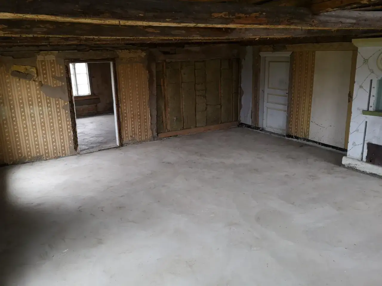 Rum under renovering med nygjutet betonggolv. V&auml;ggarna har gammal tapet och synlig isolering. Planerar att behandla betonggolv inomhus f&ouml;r en slitstark yta.