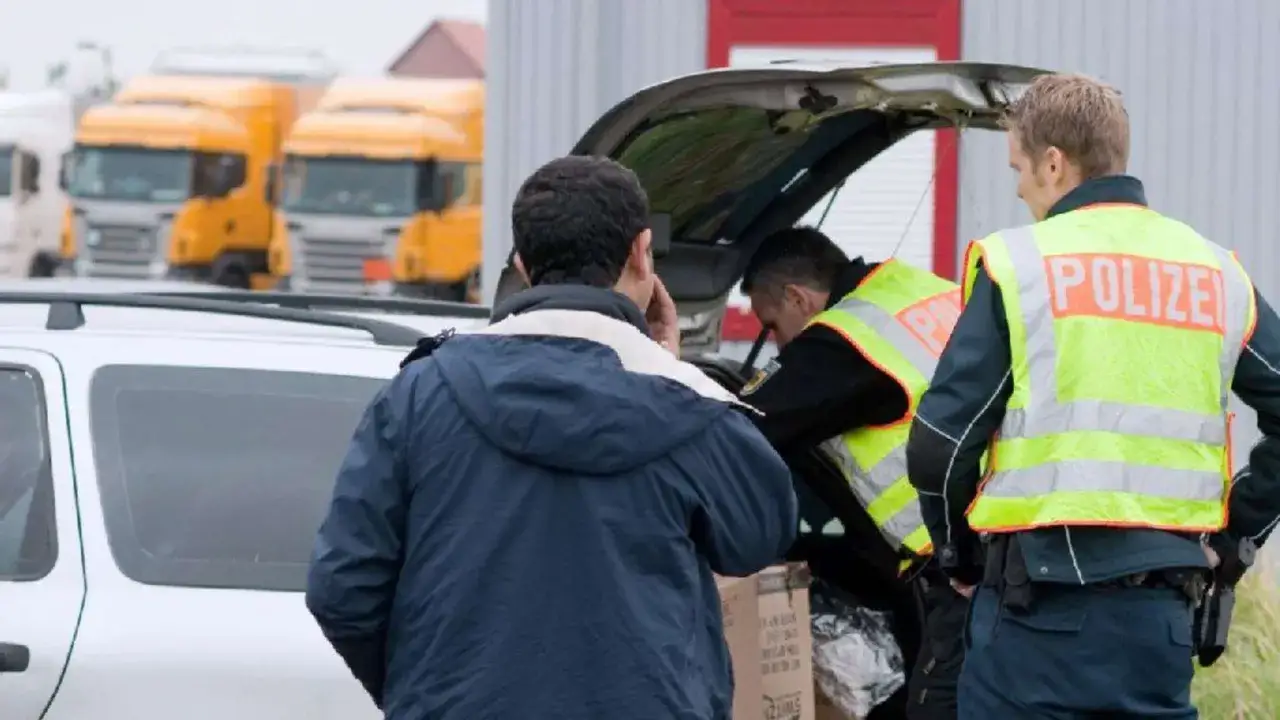 Polizisten durchsuchen ein Auto. Ein Mann telefoniert, w&auml;hrend die Beamten den Kofferraum inspizieren.