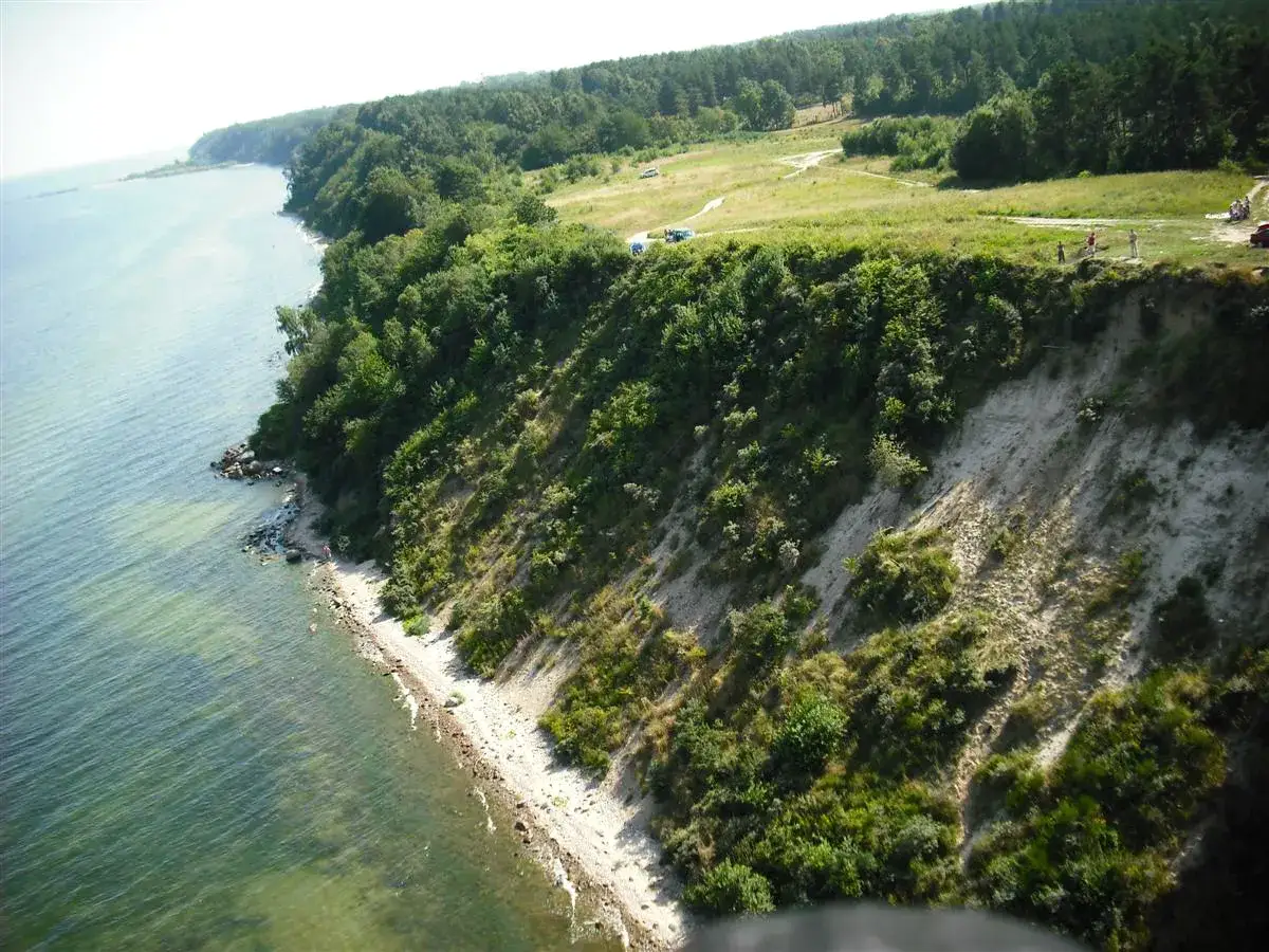 Klif w Mechelinkach, z zielonymi drzewami i morzem. W oddali widać ścieżki i ludzi, to świetne mechelinki atrakcje.