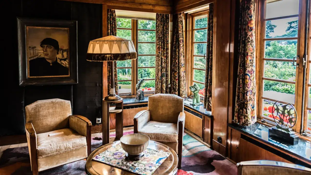 Salón con sillones de terciopelo, lámpara de pie y un retrato. La decoración evoca estilos art deco y art nouveau, con cortinas estampadas y vistas al jardín.