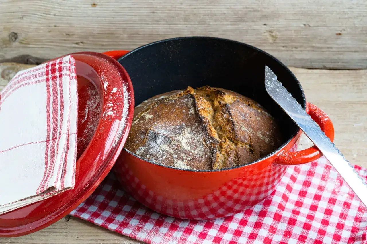 Frisch gebackenes Brot liegt in einem roten Gusseisentopf. Der Deckel mit kariertem Tuch daneben zeigt: Brot kommt nicht aus Topf, sondern aus dem Ofen.