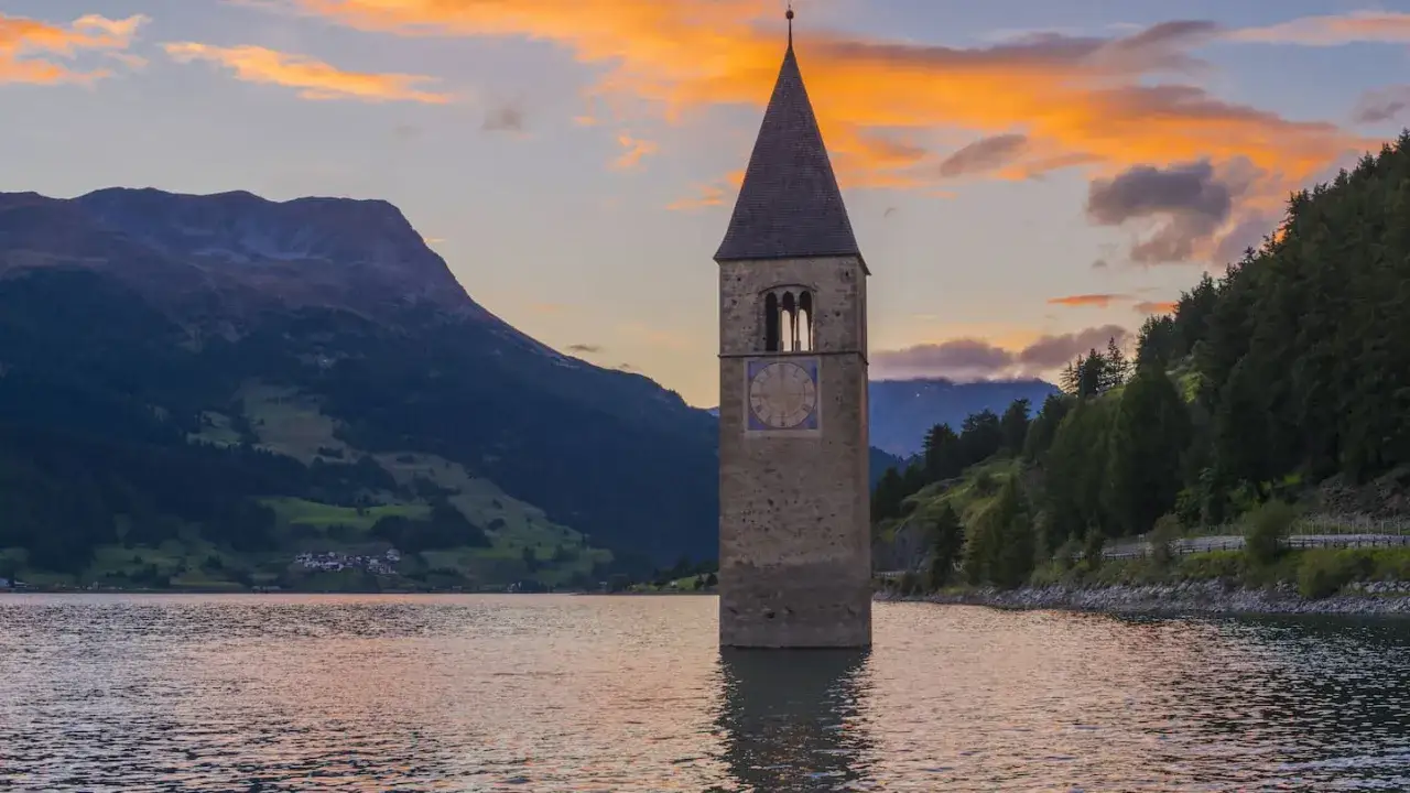 Ein Kirchturm ragt aus einem Stausee, umgeben von Bergen und Bäumen. Der Himmel ist in warmen Orange- und Rosatönen gefärbt.